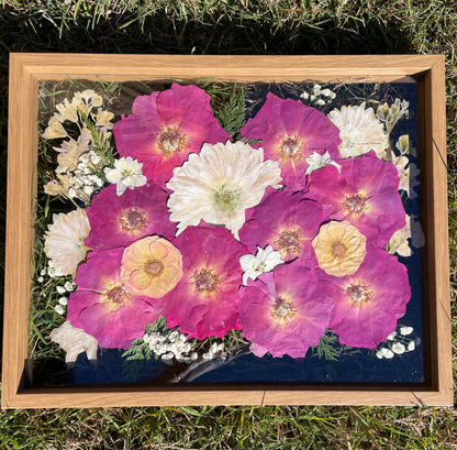 Framed artwork with pressed flowers including pink and white blossoms on a dark background.